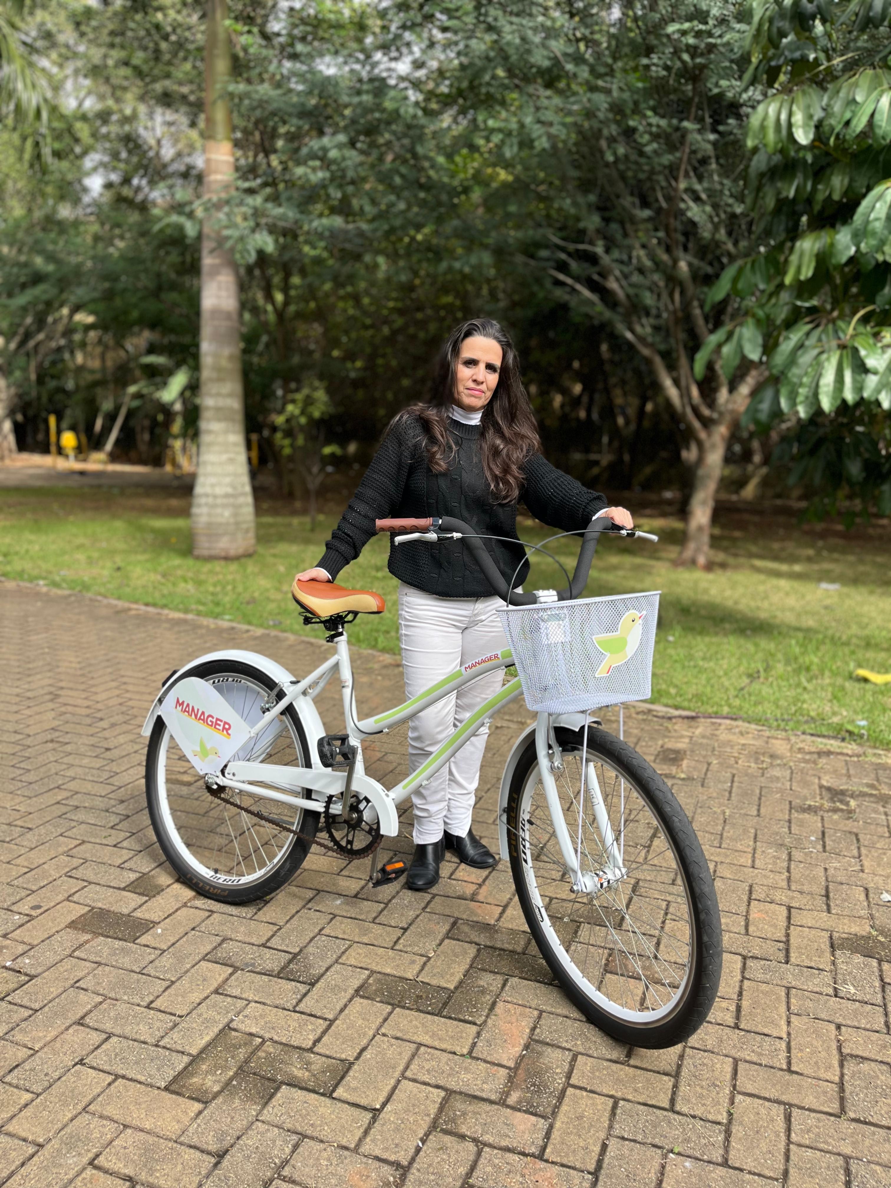 Mulher parada ao lado de bicicleta branca da Manager em área arborizada de condomínio.
